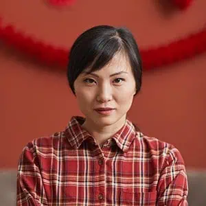 A woman with short black hair wearing a red plaid shirt, standing against a red background.